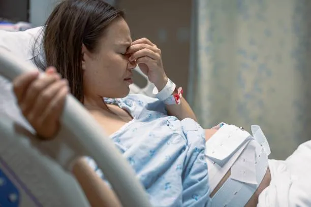 Certain Maternal or Pregnancy Conditions - drpallavichauhan Woman in a hospital bed clutching her forehead, wearing a hospital gown with a bandaged abdomen and patient bracelet on her wrist.