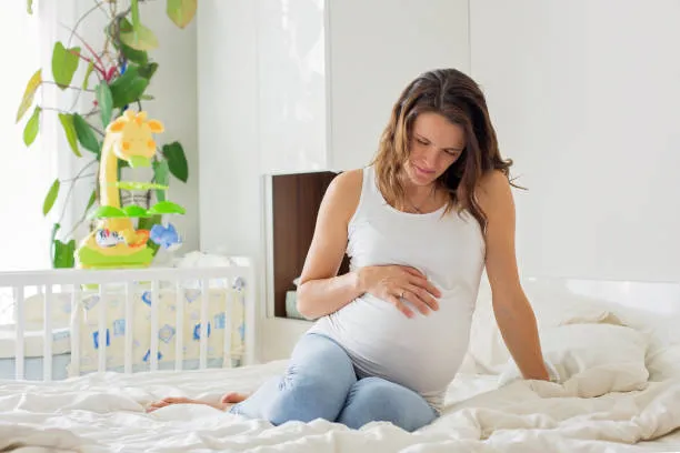 Fetal Distress - drpallavichauhan Pregnant woman sits on a bed in a bright room, cradling her baby bump with one hand.
